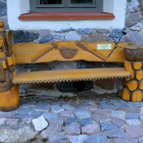 Kokgriezuma soliņš pie Sedas ūdensdzirnavu akmens sienas ar dekoratīvu figūru Kokgriezuma soliņš pie Sedas ūdensdzirnavu akmens sienas ar dekoratīvu figūru