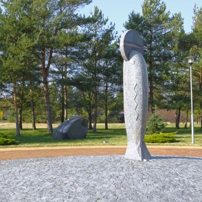 Vertikāla akmens skulptūra parkā ar priedēm un bruģētu laukumu Vertikāla akmens skulptūra parkā ar priedēm un bruģētu laukumu